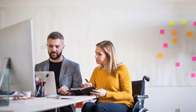 Dau berson yn eistedd wrth ddesg yn edrych ar gyfrifiadur. Mae un ohonyn nhw'n defnyddio cadair olwyn.