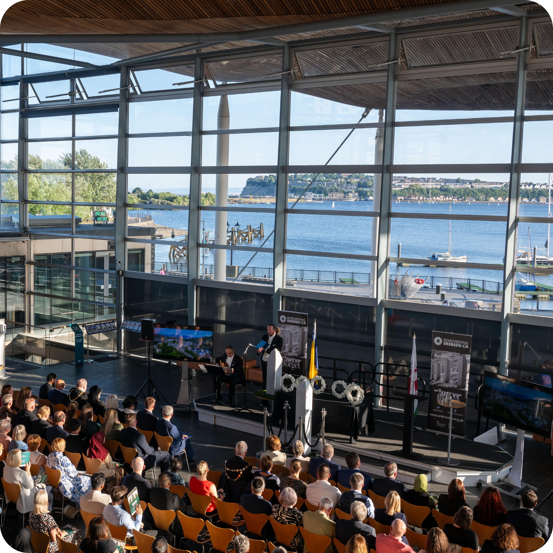Image shows visitors seated at an event in the Senedd, facing the front window while a speaker is on the stage.