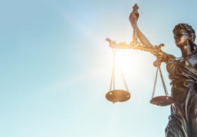 Cerflun efydd o Lady Justice yn dal clorian gytbwys yn ei llaw chwith a chleddyf yn ei llaw dde, gyda golau haul yn disgleirio trwy’r clorian yn erbyn awyr las glir.