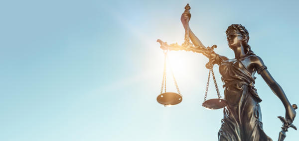 Cerflun efydd o Lady Justice yn dal clorian gytbwys yn ei llaw chwith a chleddyf yn ei llaw dde, gyda golau haul yn disgleirio trwy’r clorian yn erbyn awyr las glir.