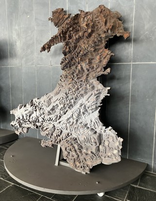 Map rhyddhad o Gymru wedi'i wneud o ddur, yn dangos tirwedd y wlad drwy haenau o linellau cyfuchlin. Mae'r map wedi'i osod ar stand metel..