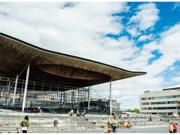 Ffotograff o du blaen adeilad y Senedd ym Mae Caerdydd. Mae'r adeilad wedi'i wneud o frics llwyd, ac mae rhai trawstiau gwyn i'w gweld. Mae arwydd sy'n dweud 'Senedd Cymru'. Yn y blaendir, mae cerddwyr a beicwyr yn mynd heibio. Mae rhai pobl yn eistedd ar y grisiau y tu allan i adeilad y Senedd.