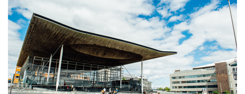 Ffotograff o du blaen adeilad y Senedd ym Mae Caerdydd. Mae'r adeilad wedi'i wneud o frics llwyd, ac mae rhai trawstiau gwyn i'w gweld. Mae arwydd sy'n dweud 'Senedd Cymru'. Yn y blaendir, mae cerddwyr a beicwyr yn mynd heibio. Mae rhai pobl yn eistedd ar y grisiau y tu allan i adeilad y Senedd.