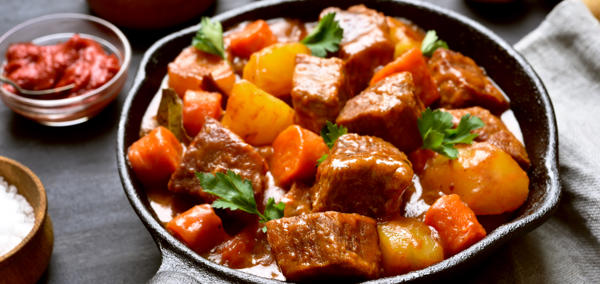 A picture of a warm meat and vegetable stew served in a small pan surrounded by ingredients. 