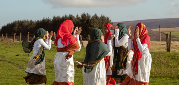 Grŵp o ferched ifanc yn gwisgo dillad traddodiadol a sgarffiau, yn sefyll mewn cylch yn dawnsio ar y bryniau ar ddiwrnod braf.