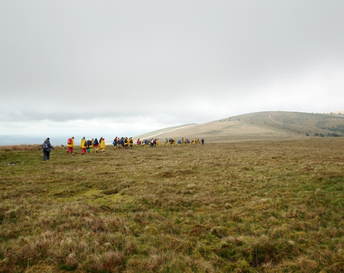 Llun sy’n dangos llecyn porfa gyda bryniau yn y cefndir. Mae grŵp o blant ysgol sy’n gwisgo cotiau glaw melyn a bagiau ar eu cefn yn cerdded tuag at y bryniau. Mae'r awyr yn gymylog a llwyd yr olwg.