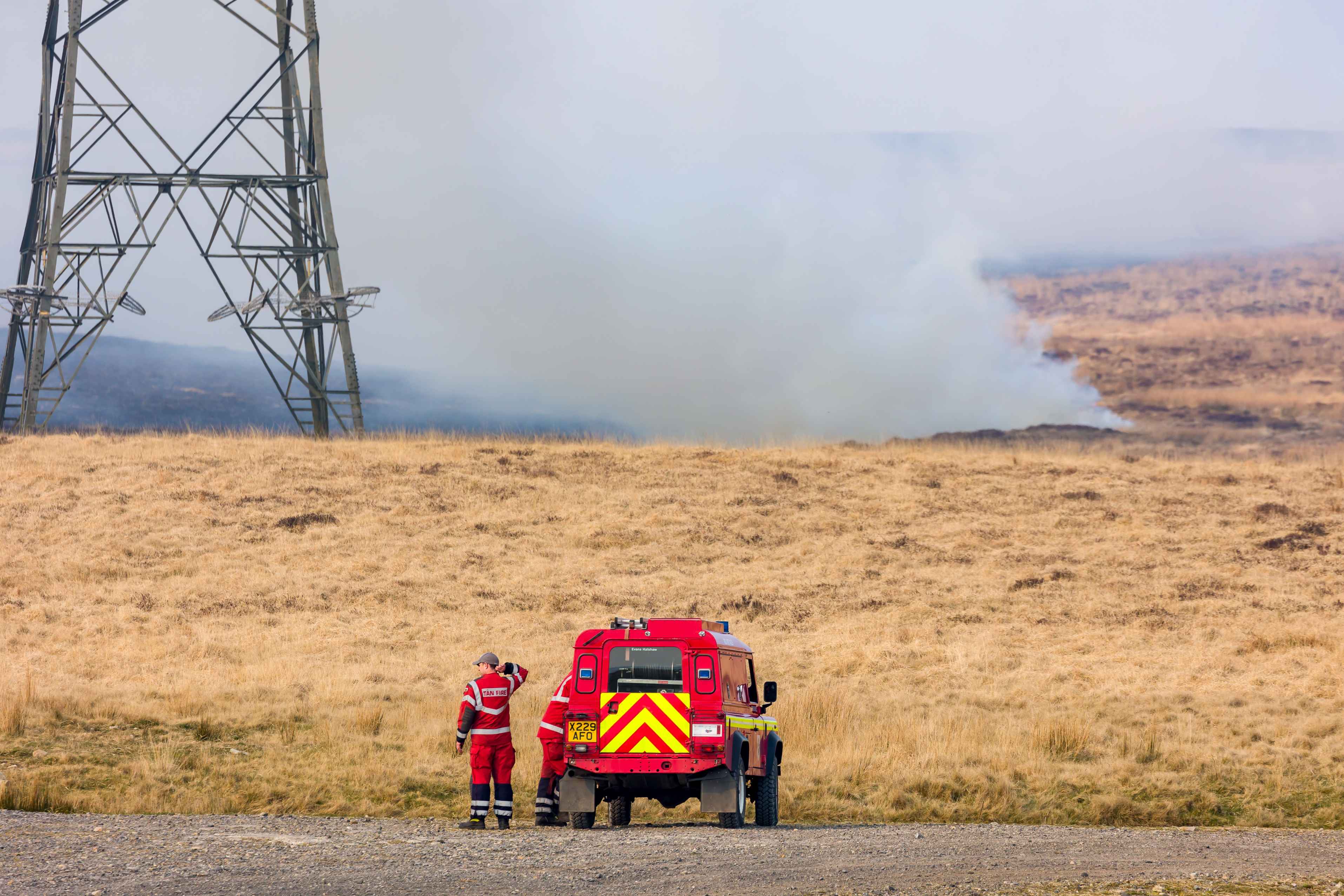 Ffotograff tirwedd o injan dân o flaen tân glaswellt ar rostir, gyda mwg yn codi tuag at beilon yn y cefndir.