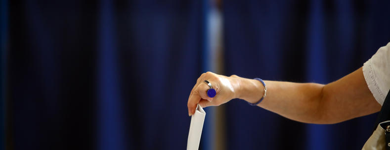 Braich fenywaidd yn gosod papur pleidleisio mewn blwch pleidleisio gyda chefndir glas.