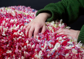 Llun agos o berson yn dal gwrthrych blewog. Defnyddiwyd gwahanol liwiau o edafedd gan gynnwys pinc, gwyrdd, coch a glas.