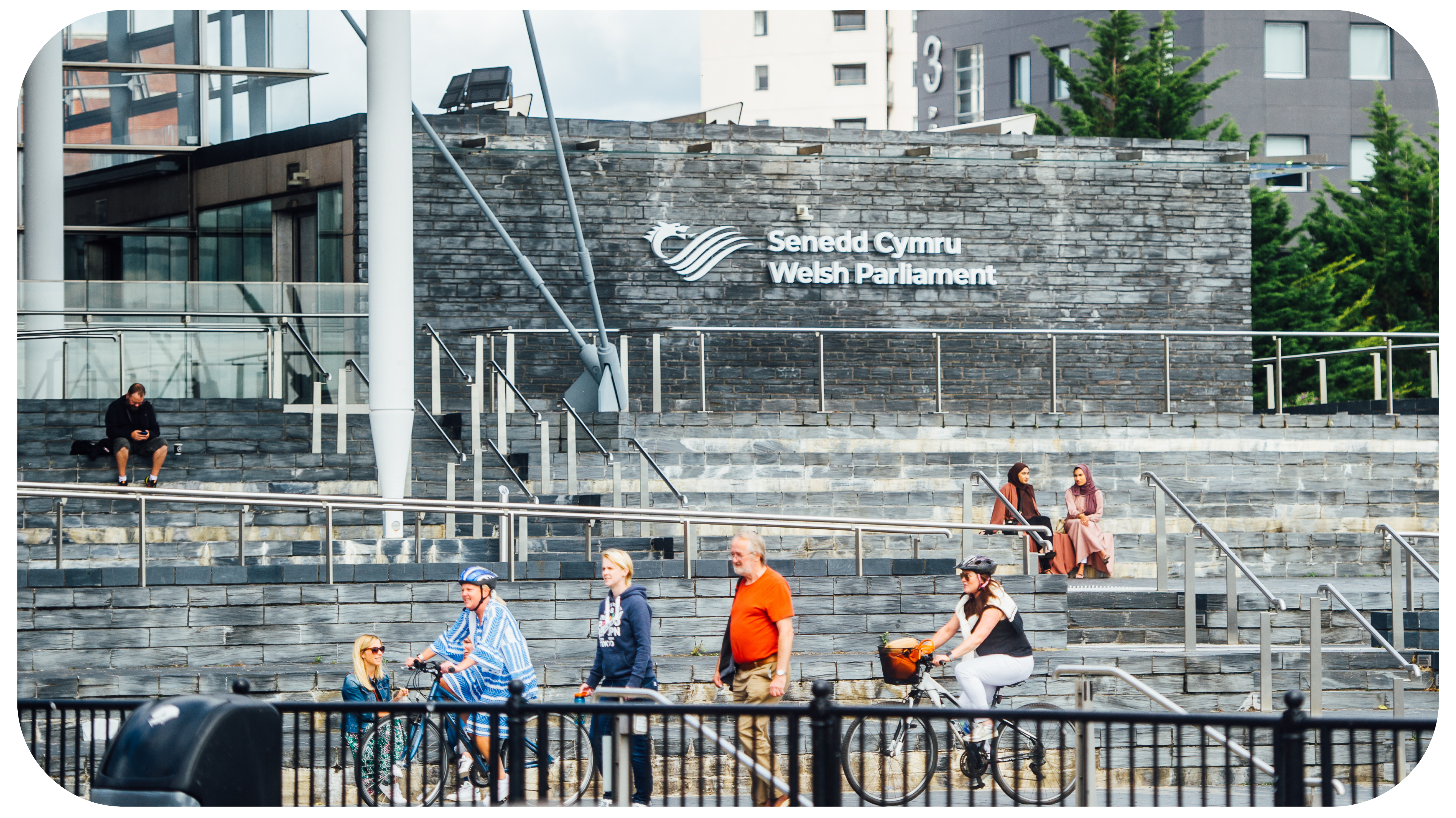 A photograph of the front of the Senedd building in Cardiff Bay. The building is comprised of grey bricks, and some white beams are visible. There is a sign that reads ‘Welsh Parliament’. In the foreground, there are pedestrians and cyclists passing by. Some people are sitting on the steps outside the Senedd building.