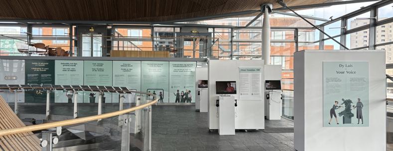 Y tu mewn i'r Senedd, yn dangos arddangosfa, gyda thestun ar fyrddau gwybodaeth a fideos ar sgriniau.