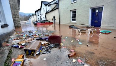 This is a picture of a street flooded by muddy water with floating rubbish and domestic items in the foreground. In the background are vehicles, most of which are submerged, one is  There is one floating car.