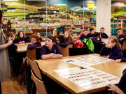 Mae athrawes yn siarad â dosbarth o blant oedran cynradd. Mae'r plant yn gwrando, yn wynebu ymlaen. Mae llun mawr ar y wal o dirweddau cymysg trefol a gwledig.